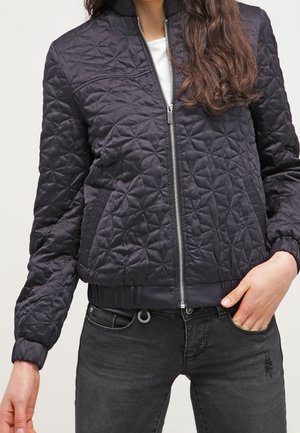 Frau trägt eine schwarze gesteppte Jacke mit Reißverschluss, mit einem gerippten Kragen und Bündchen, kombiniert mit schwarzen Jeans, eine Hand in der Tasche.