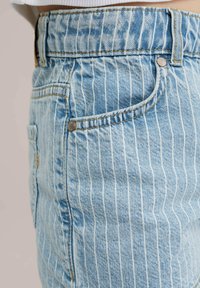 Svijetloplave traperice s vertikalnim bijelim prugama, koje imaju bočnu džepu, bakrenu zakovicu i vidljive detalje šivanja.