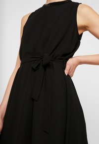 Robe noire sans manches avec un col orné de perles et une ceinture nouée à la taille, portée par une personne avec une main sur la hanche.