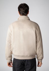 Beige Wildlederjacke mit hohem Kragen, gerippten Bündchen und Taillenbund. Besitzt eine glatte Textur, einen lässigen Schnitt und keine sichtbaren Metallteile.
