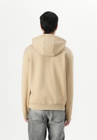 Sweatshirt bege com capuz e um ajuste relaxado, com punhos e barra canelados. O material parece suave e texturizado, combinado com jeans cinzentos claros.
