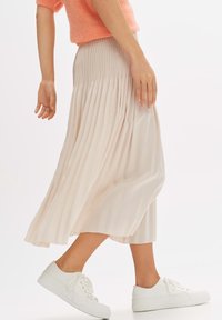 Gelaagde midi-rok in zacht beige met een geribbelde tailleband, gecombineerd met witte sneakers. De stof heeft een gladde, vloeiende textuur.