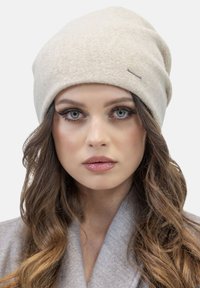 Beige gebreide beanie met een zachte textuur, gekenmerkt door een ontspannen pasvorm en een subtiele logo-accent aan de zijkant.