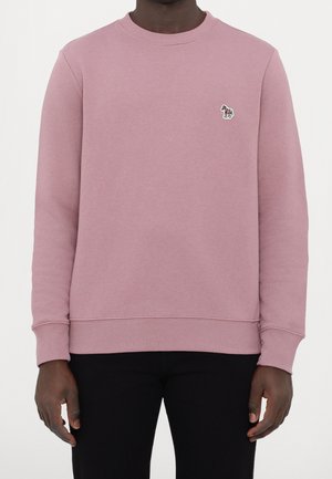 Homme portant un sweat-shirt rose poudré à col rond avec un petit logo zèbre multicolore sur la poitrine gauche et un pantalon noir.