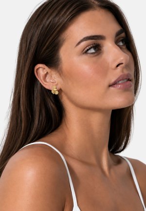 Femme aux longs cheveux foncés portant un débardeur blanc et de petites boucles d’oreilles en grappes de disques dorés, regardant légèrement vers le haut à droite.