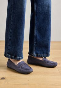Lilla nahast loafers, millel on õmmeldud õmblusdetail ja lame kummitald, kantud laia säärega siniste teksadega, näidates juhuslikku stiili puidust põrandal.