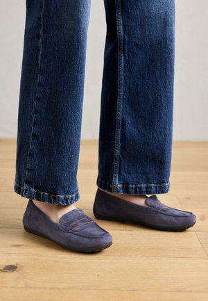 Paarse suede instappers met een gestikt naaddetail en een platte rubberen zool, gedragen met wijde blauwe jeans, die een casual stijl op een houten vloer tonen.