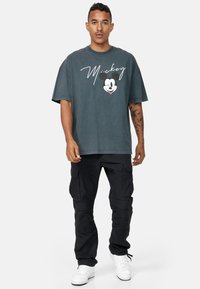 Camiseta gris de gran tamaño con un gráfico en blanco y negro de Mickey Mouse y texto en cursiva blanca. Combinada con pantalones cargo negros y zapatillas blancas.