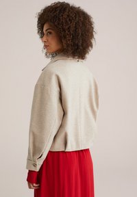 Beige, tekstureret uld-blandingsjakke med klassisk krave, oversized pasform og opflettede ærmer. Båret over en flydende rød kjole.