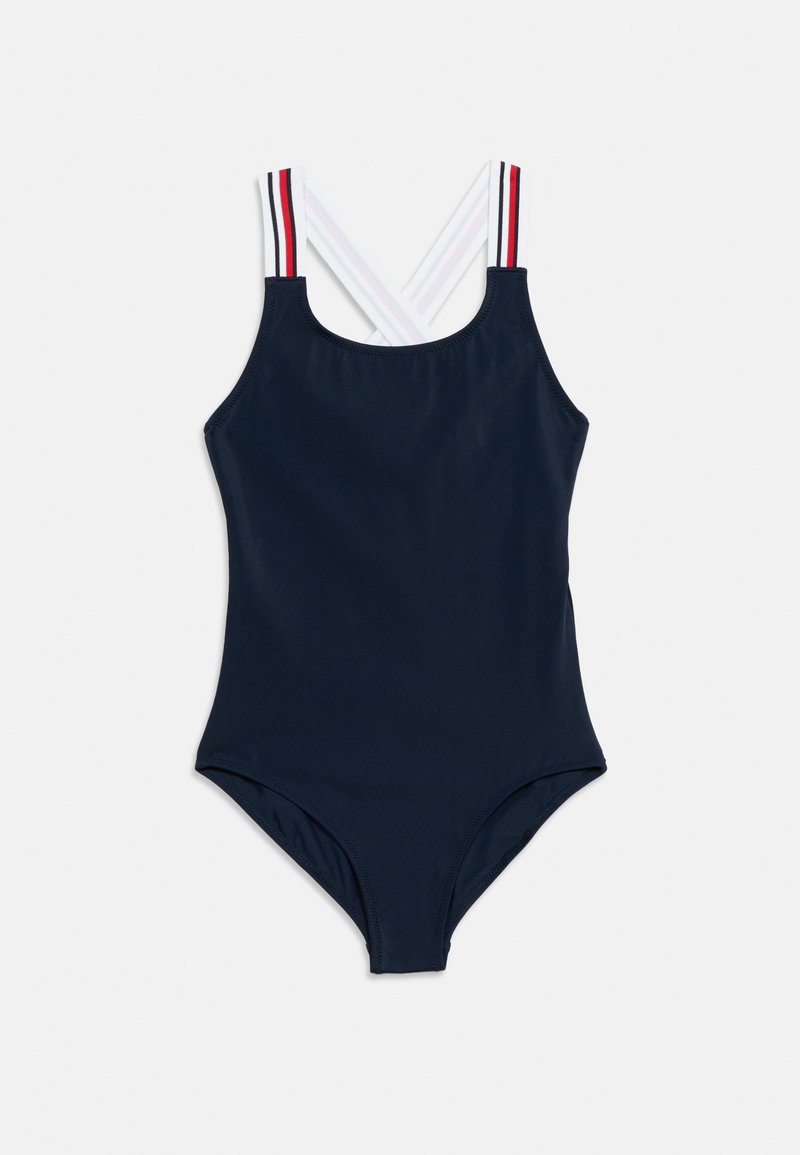 Ολόσωμο μαγιό σε ναυτικό μπλε χρώμα με λευκές τιράντες στους ώμους που διασταυρώνονται στην πλάτη, με κόκκινες και μαύρες ρίγες στις εξωτερικές άκρες.