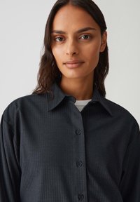 Donna con pelle di media tonalità marrone e lunghi capelli scuri, che indossa una camicia scura con bottoni e una maglietta bianca, contro uno sfondo chiaro e semplice.