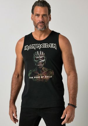 Mężczyzna w czarnym podkoszulku Iron Maiden "The Book of Souls", stojący na jednolitym jasno-szarym tle, patrzący przed siebie.