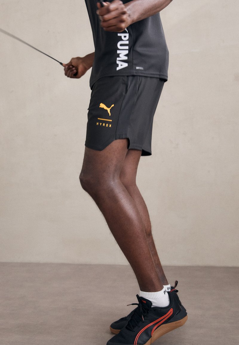 Zwarte sportshorts met een gebogen zoom en een oranje Puma-logo, gecombineerd met zwarte sneakers met rode accenten en witte sokken.
