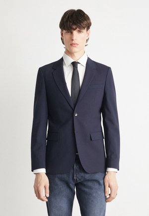 Blazer blu navy realizzato in tessuto liscio, vestibilità slim, chiusura a un bottone, revers a lancia, due tasche frontali, indossato sopra una camicia bianca e una cravatta nera.