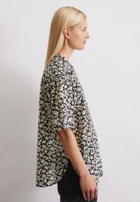 Chemise à manches courtes noire avec un motif de feuilles blanches, présentant une coupe ample et un ourlet arrondi, fabriquée en tissu léger.