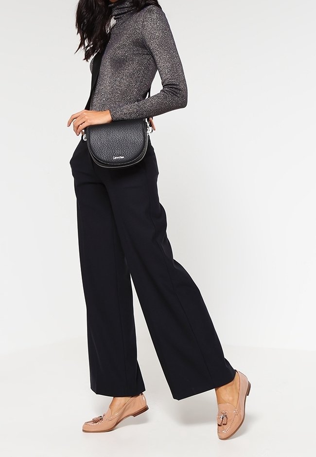 Czarna torebka na ramię o teksturowanej powierzchni, zestawiona z ciemnym golfem i czarnymi, szerokimi spodniami. Jasnobrązowe mokasyny z dekoracyjnymi detalami.