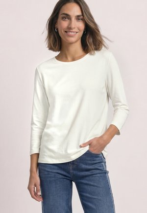 Kvinde smilende, iført en ensfarvet hvid langærmet skjorte og blå jeans, med den ene hånd i lommen mod en neutral baggrund.