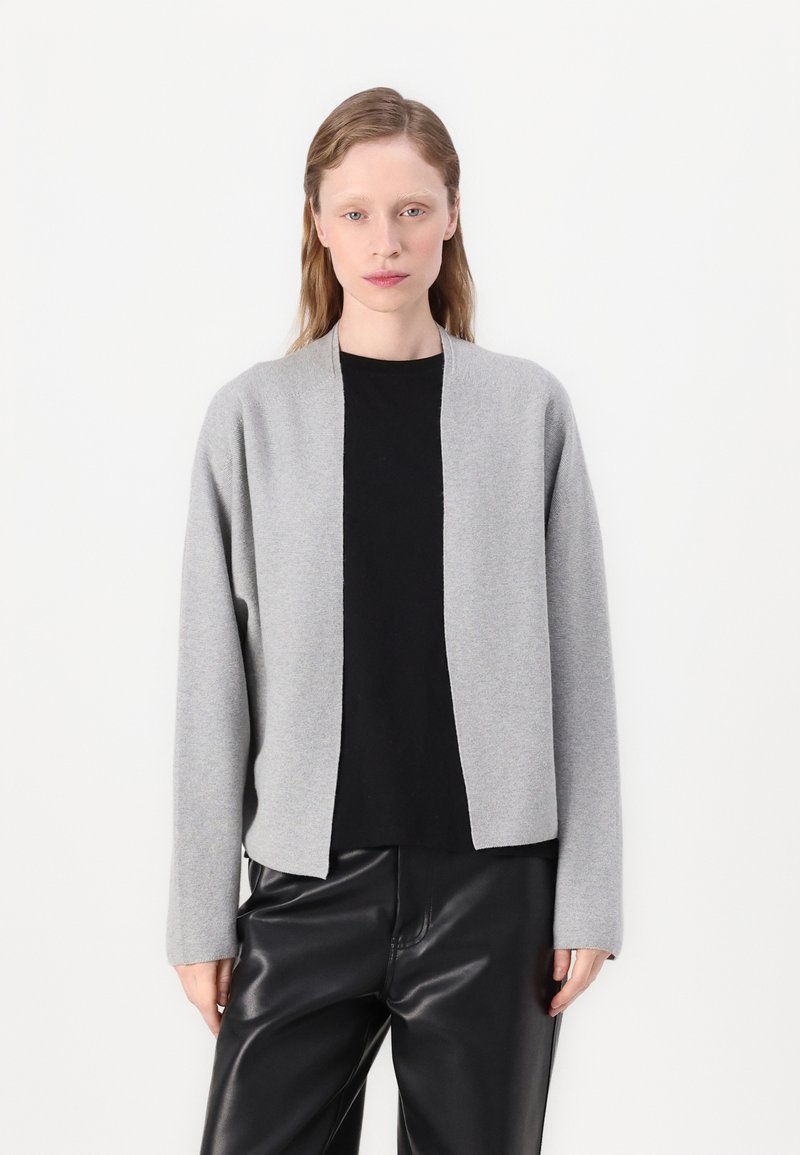 Šedý pletený cardigan s otevřeným předním dílem a dlouhými rukávy, je vrstvený přes černý top. Hladká textura, minimalistický design a volný střih.