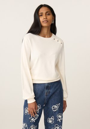 Vrouw met lang donker haar draagt een witte trui met drie knopen op de schouder en blauwe spijkerbroek met witte bloemrijke borduursels.