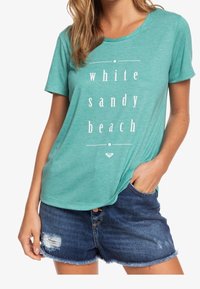 T-shirt teal a maniche corte con testo bianco "spiaggia di sabbia bianca", abbinato a pantaloni corti di denim logori. Fit casual, morbido e leggero.
