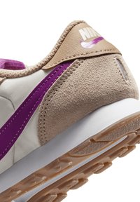 Zapatilla ligera con parte superior de ante beige y textil blanco, acentos morados y suela de goma texturizada. Presenta un logo prominente en el talón.