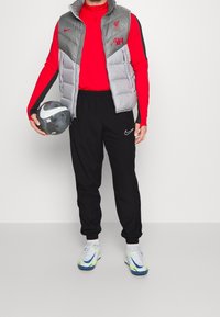 Grijze gewatteerde vest over een rood longsleeve shirt, zwarte broek en multicolore Nike sneakers; houd een zwart-wit voetbal vast.