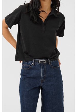 Vrouw die een zwart poloshirt met korte mouwen draagt, ingestopt in een hooggetailleerde blauwe spijkerbroek met een zwarte riem, handen op de heupen.