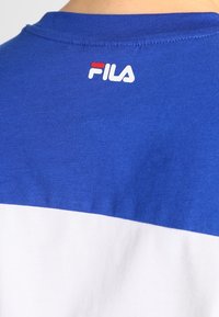 Blå och vit skjorta bakifrån som visar FILA-logotypen i vitt och rött nära halslinjen.