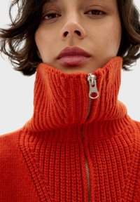 Czerwony sweter z dzianiny z wysokim kołnierzem i zamkiem błyskawicznym z przodu. Teksturowany wzór tworzy głębię, z prążkowanym designem na kołnierzu i rękawach.