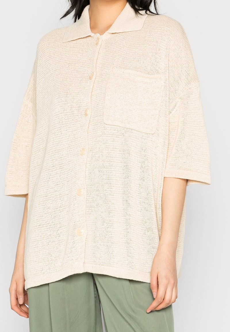 Femme portant une chemise en maille crème ample à manches courtes avec col et poche poitrine, associée à un pantalon vert clair.