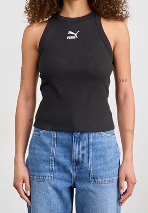 Kobieta ubrana w czarny bezrękawnik Puma z prążkowanej tkaniny i niebieskie jeansy z wysokim stanem oraz przednimi naszywanymi kieszeniami, stoi na jednolitym tle.