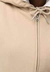 Haut à capuche zipé beige en tissu doux avec une fermeture éclair à l'avant et un cordon de serrage pour la capuche. Présente une texture lisse et un design minimal.