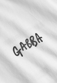 Țesătură de bumbac albă cu o textură moale. Cuvântul "GABBA" este prezentat proeminent cu litere negre îngroșate.