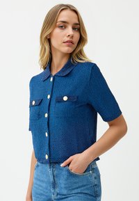 Junge Frau mit blonden Haaren, die eine strukturierte blaue Kurzarmjacke mit goldenen Knöpfen und blaue Jeans trägt, mit der Hand in der Tasche.