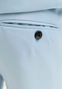 Close-up van lichtblauwe broek met een achterzak met bies, bevestigd met een zwarte knoop.