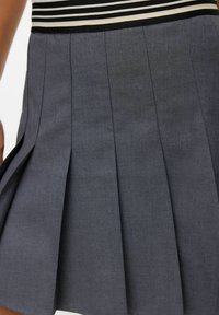 Gros plan sur une jupe plissée grise avec une taille rayée noir et blanc, portée par une personne sur fond blanc.