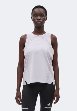 Femeie purtând un top sport alb fără mâneci de la Adidas și colanți negri cu logo-uri Adidas, stând în fața unui fundal simplu și deschis.
