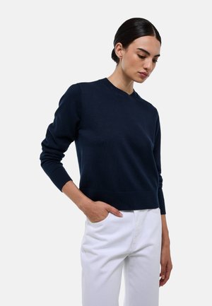 Pull en tricot bleu marine avec un col rond et des poignets côtelés, associé à un pantalon blanc, montrant une silhouette cintrée.
