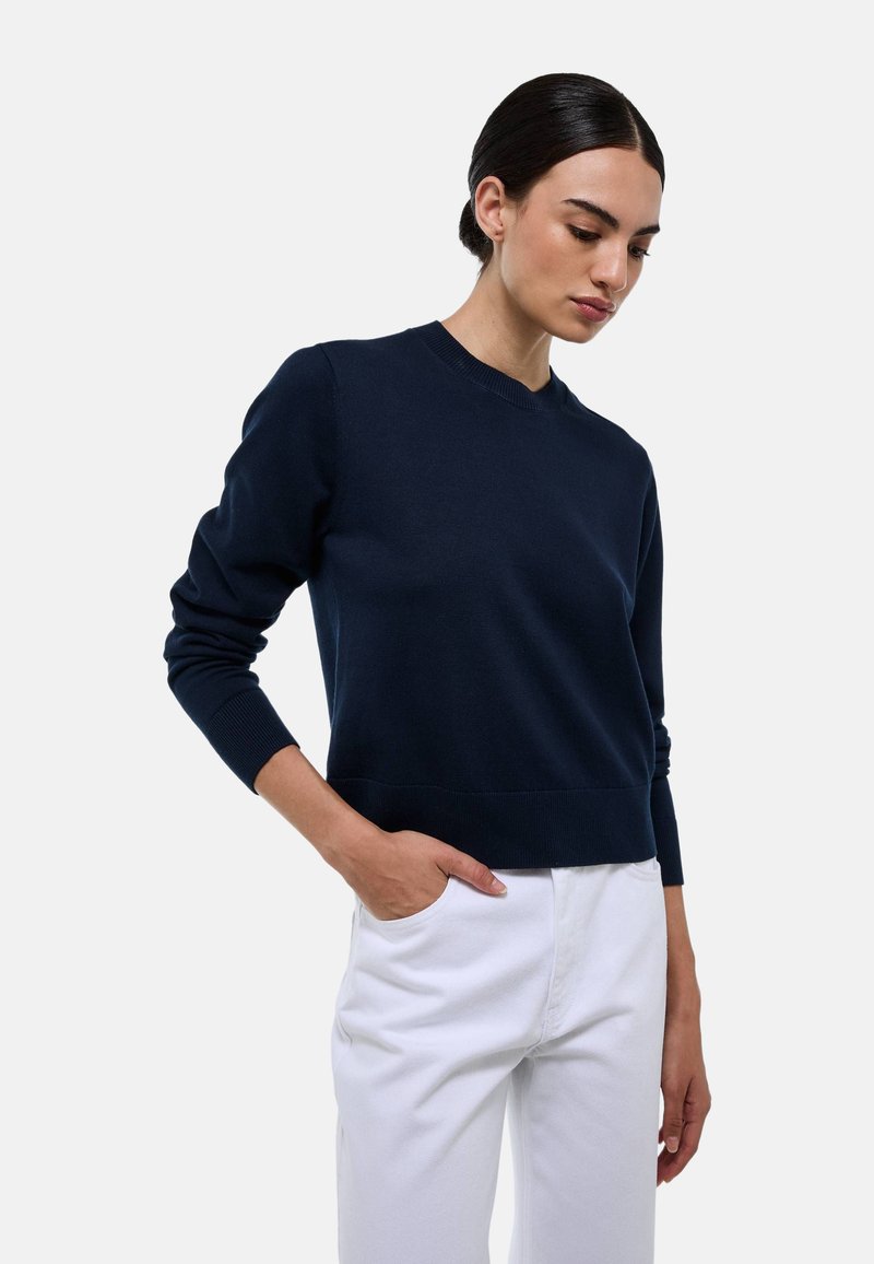 Pull en tricot bleu marine avec un col rond et des poignets côtelés, associé à un pantalon blanc, montrant une silhouette cintrée.
