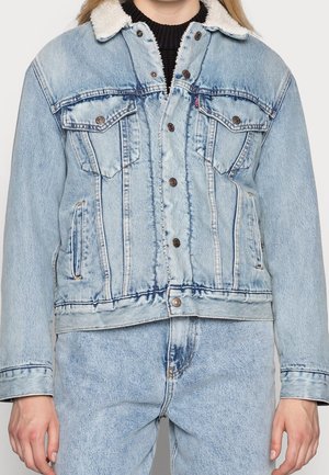 Personne portant une veste en jean bleu clair avec un col sherpa blanc et un jean en denim bleu clair assorti, debout devant un fond uni.