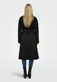Long manteau en laine noir avec une taille ceinturée, manches longues et une longueur sous le genou, associé à un jean skinny bleu et des bottes noires à talons.