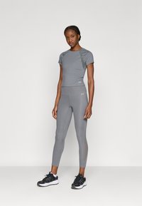 Siva sportska odjeća koja uključuje majicu kratkih rukava i uske tajice. Oba dijela imaju glatku teksturu i suptilne naglašene detalje. Crne tenisice upotpunjuju izgled.