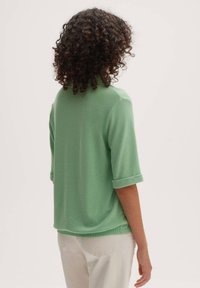 Zelený pulovr s polovinou rukávů, volnějšího střihu, vyrobený z měkké tkaniny, s ohrnutými manžetami a jemnou texturou.