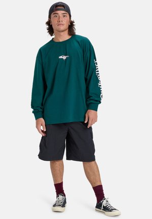 Långärmad teal skjorta med en liten logotyp på bröstet och vit text på ärmen, kombinerad med svarta cargoshorts och sneakers.