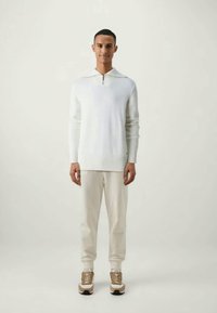 Λευκό πλεκτό πουλόβερ με ριγωτό στρίφωμα και γιακά, συνδυασμένο με ανοιχτό μπεζ φόρμες και μπεζ αθλητικά παπούτσια. Απλός, μοντέρνος σχεδιασμός.