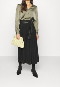 Blouse en satin vert olive, jupe midi plissée noire, ceinture noire avec détail nœud, sac à main tissé jaune et chaussures noires à bout pointu.