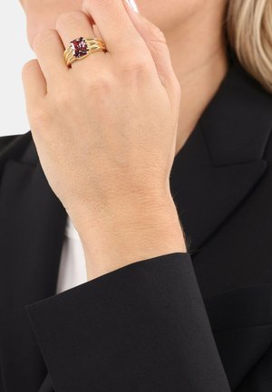 Ring in Goldoptik mit großem rotem Edelstein, bestehend aus vier schlanken Ringen, getragen an einer Hand vor dem Hintergrund einer schwarzen Jacke.