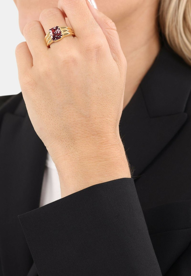 Anillo tono dorado con una gran piedra preciosa roja, con cuatro bandas delgadas, llevado en una mano contra un fondo de chaqueta negra.