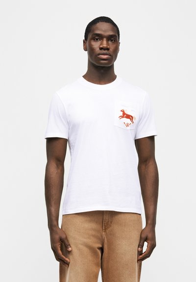 Άνδρας που φοράει λευκό κοντομάνικο μπλουζάκι με κέντημα ενός αλόγου στην αριστερή τσέπη του στήθους και ανοιχτό καφέ παντελόνι, στέκεται μπροστά σε απλό φόντο.