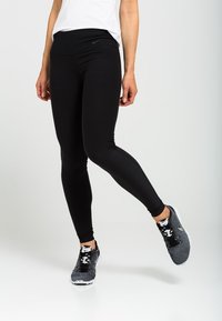 Persoon in zwarte strakke leggings en grijze Nike hardloopschoenen staat op één voet tegen een witte achtergrond.